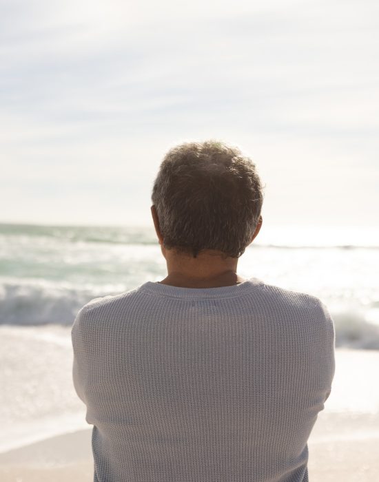Rear view of biracial senior man looking at waves crashing in sea against sky from sunny beach